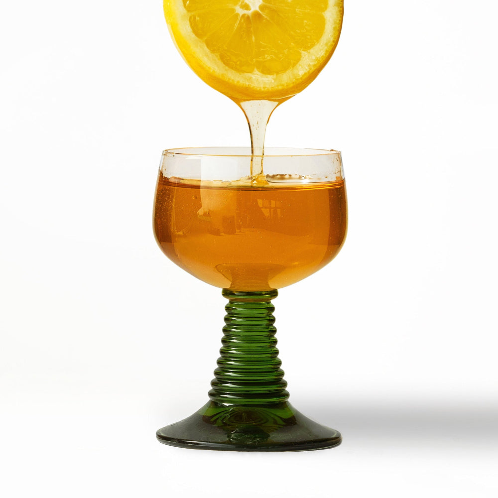 Glass with green stem being filled with honey from a lemon slice against a white background